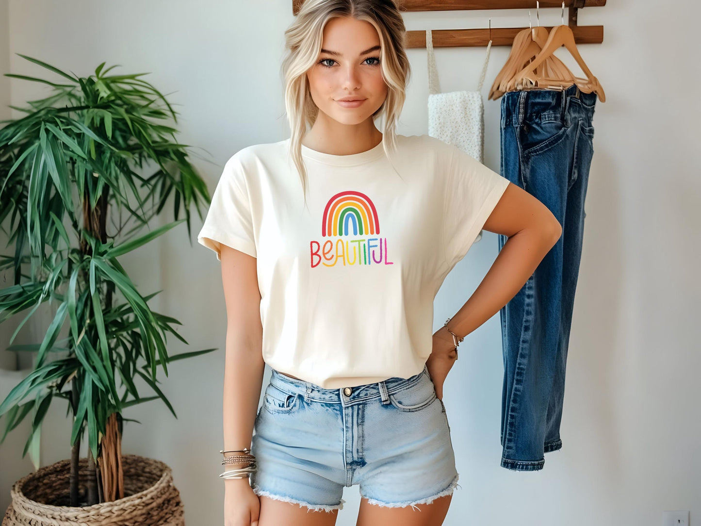 Stylish woman in a cream t-shirt with rainbow “BEAUTIFUL” text, wearing denim shorts and standing in a bright room with plants and hanging clothes.