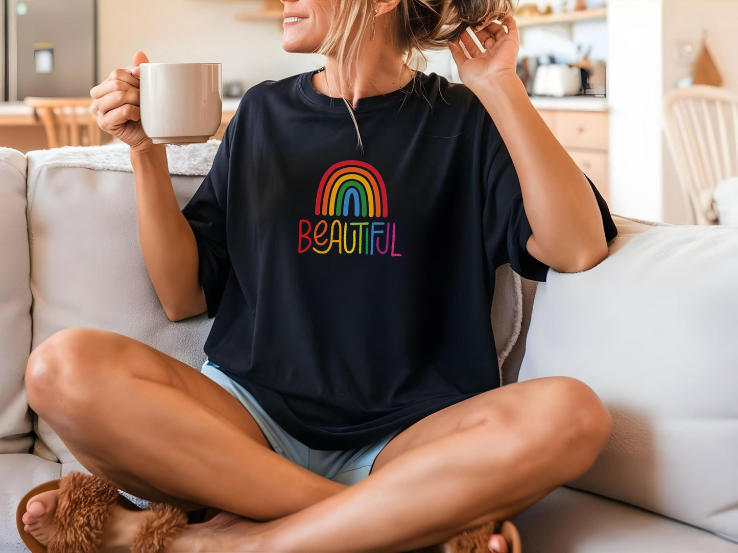 Woman relaxing on a couch wearing a black t-shirt with colorful “BEAUTIFUL” text and rainbow design, holding a mug and smiling.