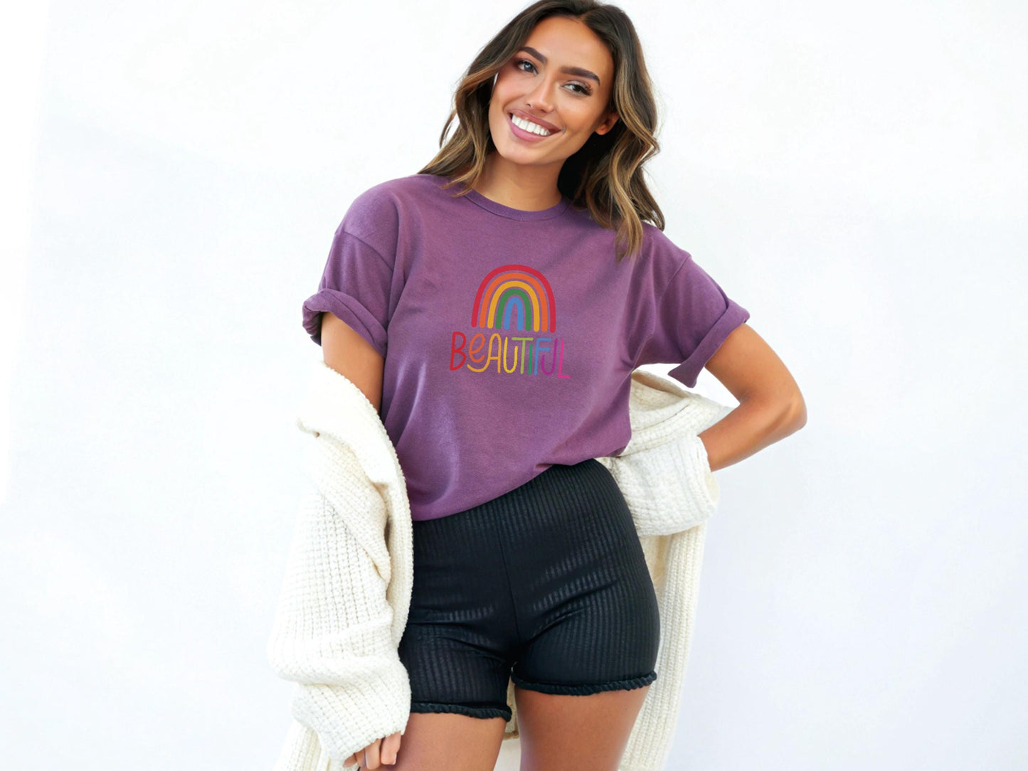 Smiling woman in a purple rolled-sleeve t-shirt featuring a rainbow and “BEAUTIFUL” design, paired with black shorts and a cozy cream cardigan.