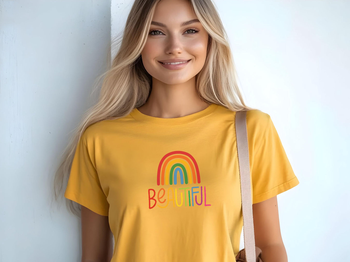 Blonde woman wearing a sunny yellow t-shirt with rainbow “BEAUTIFUL” graphic, smiling and holding a tote bag against a light wall.