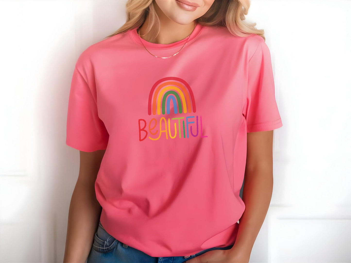 Close-up of a pink t-shirt with rainbow “BEAUTIFUL” design worn by a woman with blonde hair, posing in front of a clean white background.