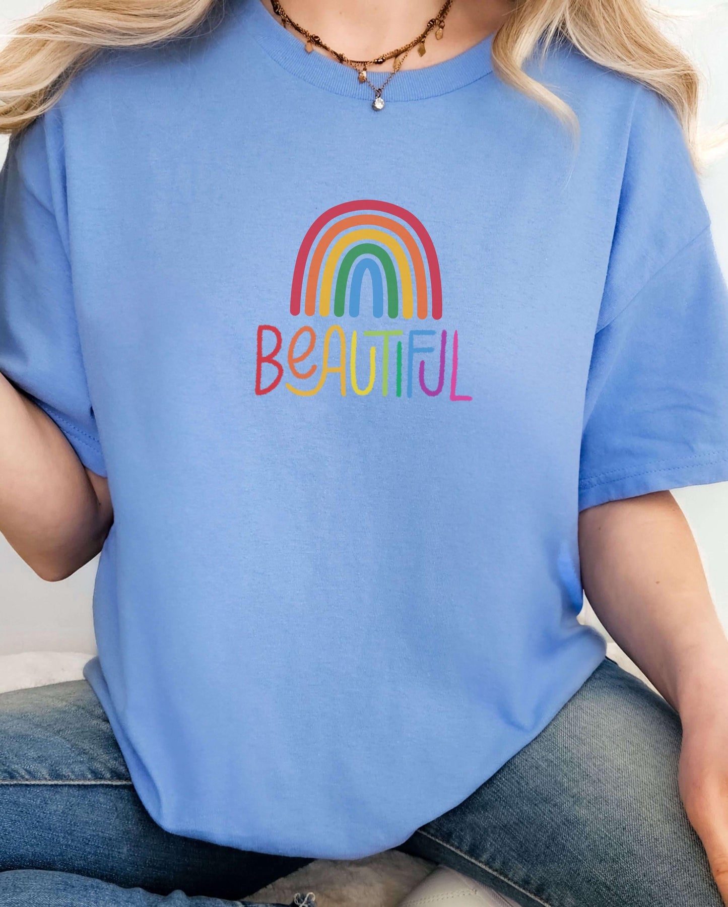 Light blue t-shirt with a rainbow and “Beautiful” printed in bright colors, worn by a woman in jeans. Positive graphic tee, mental health awareness shirt, pastel pride shirt, self-love fashion.