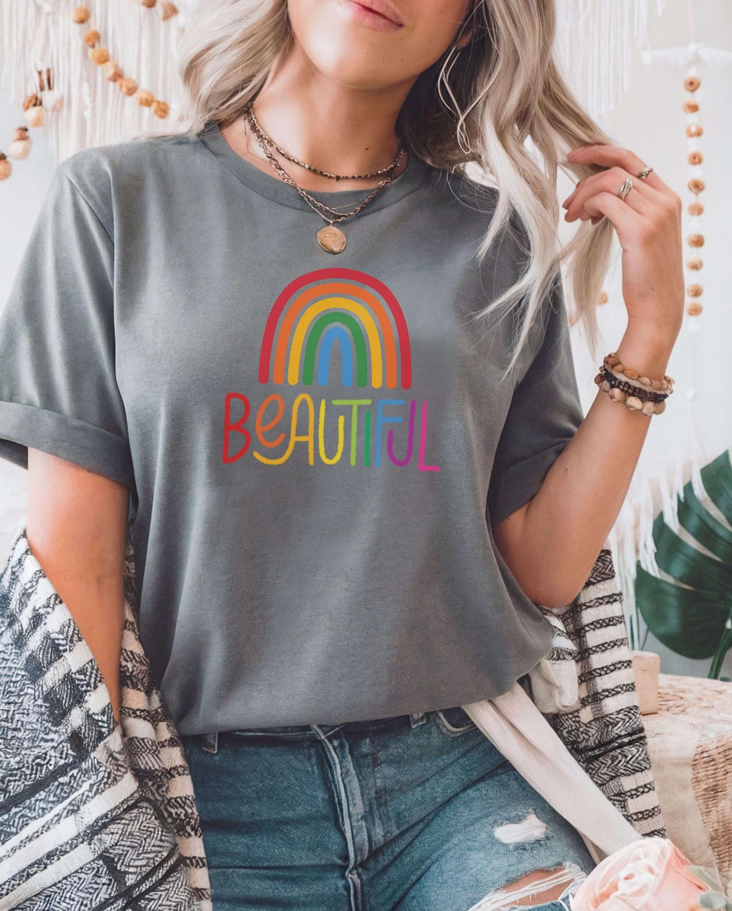 Woman in a soft gray t-shirt with rainbow “Beautiful” graphic, styled with layered necklaces and casual denim. Rainbow aesthetic shirt, modern inclusive tee, stylish mental health awareness shirt.