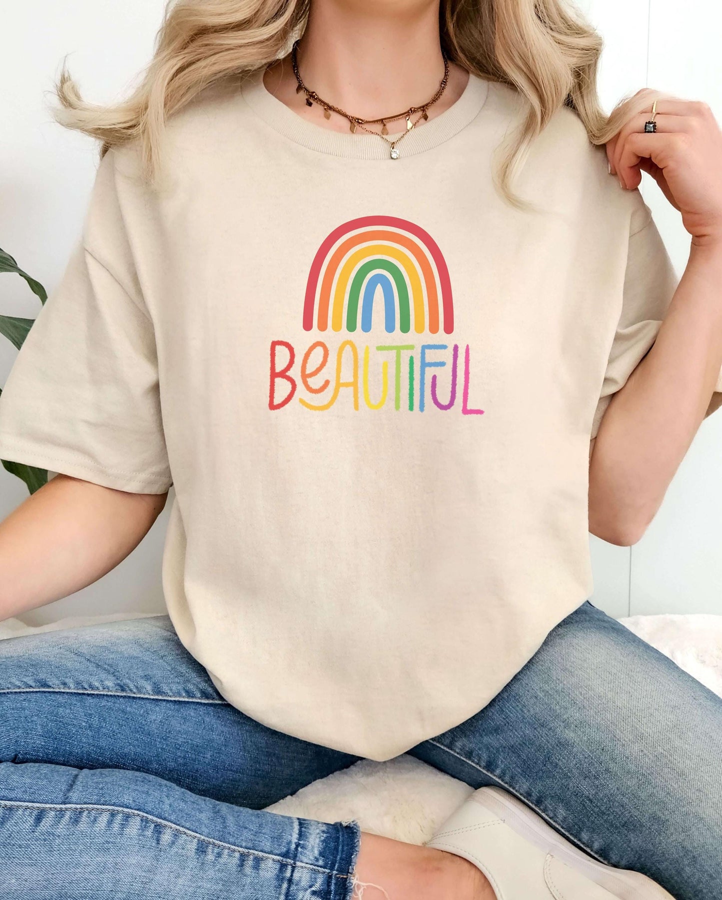 Cream beige t-shirt with rainbow “Beautiful” print, worn by woman in blue jeans. Neutral tone pride shirt, inclusive everyday wear, simple self-love tee for women.