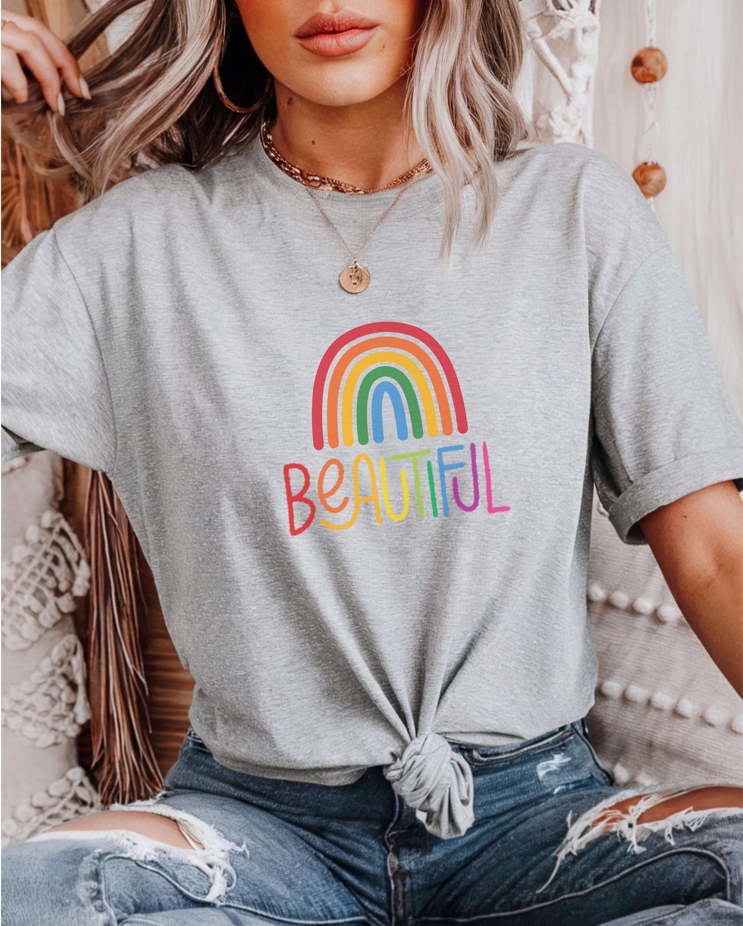 Light gray t-shirt with rainbow “Beautiful” text and rainbow graphic, styled with ripped jeans and a knotted front. Worn by a woman with wavy blonde hair and layered gold jewelry. LGBTQ+ pride shirt, positive message tee, rainbow graphic top, empowering casual outfit.