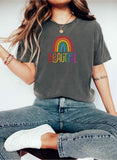 Woman sitting cross-legged on a stool wearing a faded charcoal t-shirt with a colorful rainbow and the word “BEAUTIFUL” printed in rainbow colors. She’s accessorized with a necklace, beaded bracelets, and casual jeans, creating a relaxed, boho-chic vibe.