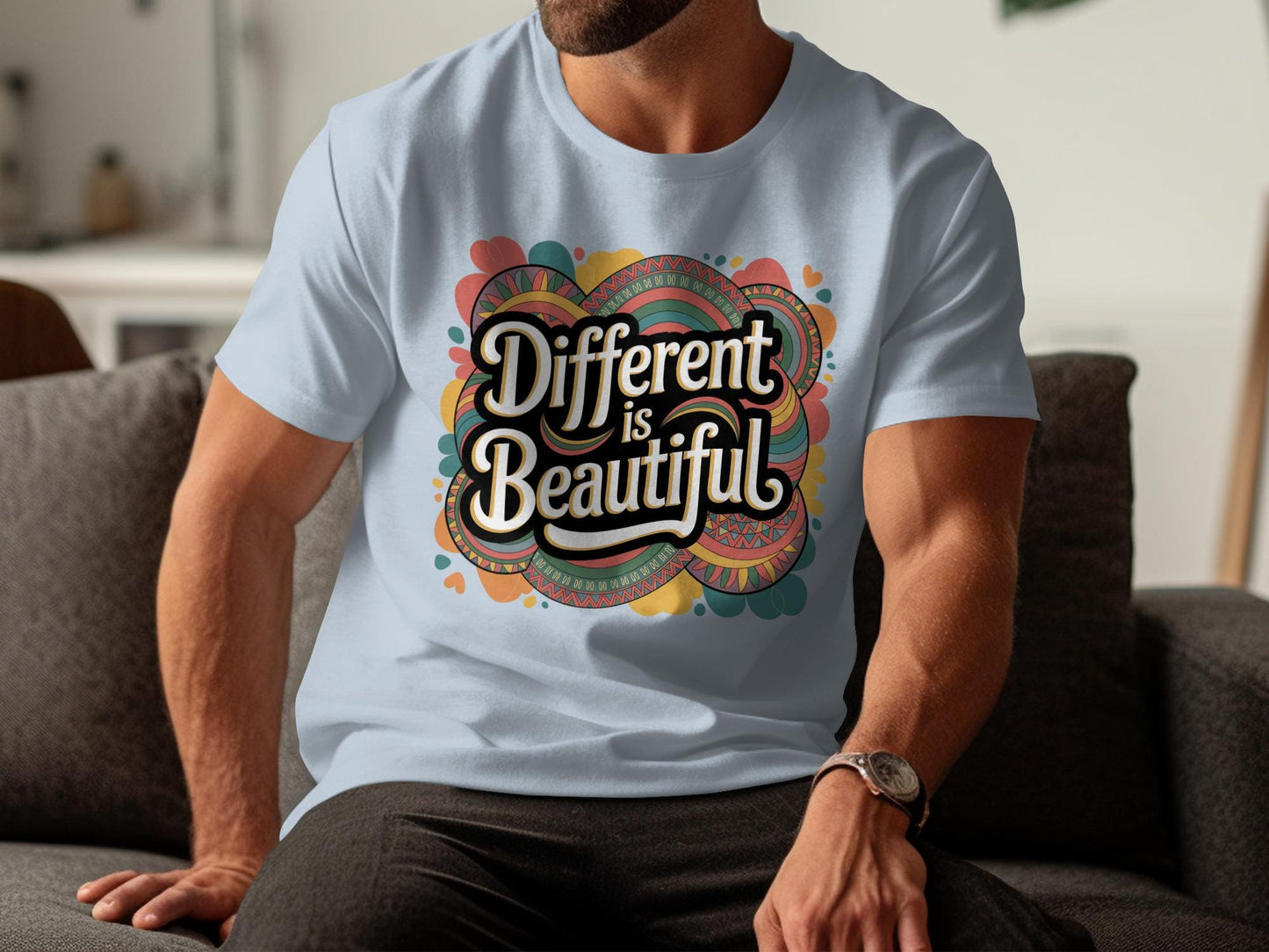 Man sitting indoors wearing a light blue t-shirt with a bold retro design that says “Different is Beautiful” in large white lettering. The colorful graphic includes hearts, swirls, and abstract patterns in teal, pink, mustard, and orange. This positive quote tee is a colorful graphic tee and custom graphic print, making it a bold shirt, emotional shirt, and unique statement tee. Perfect for anyone wanting a stand out shirt or self love t shirt with a positive message top.