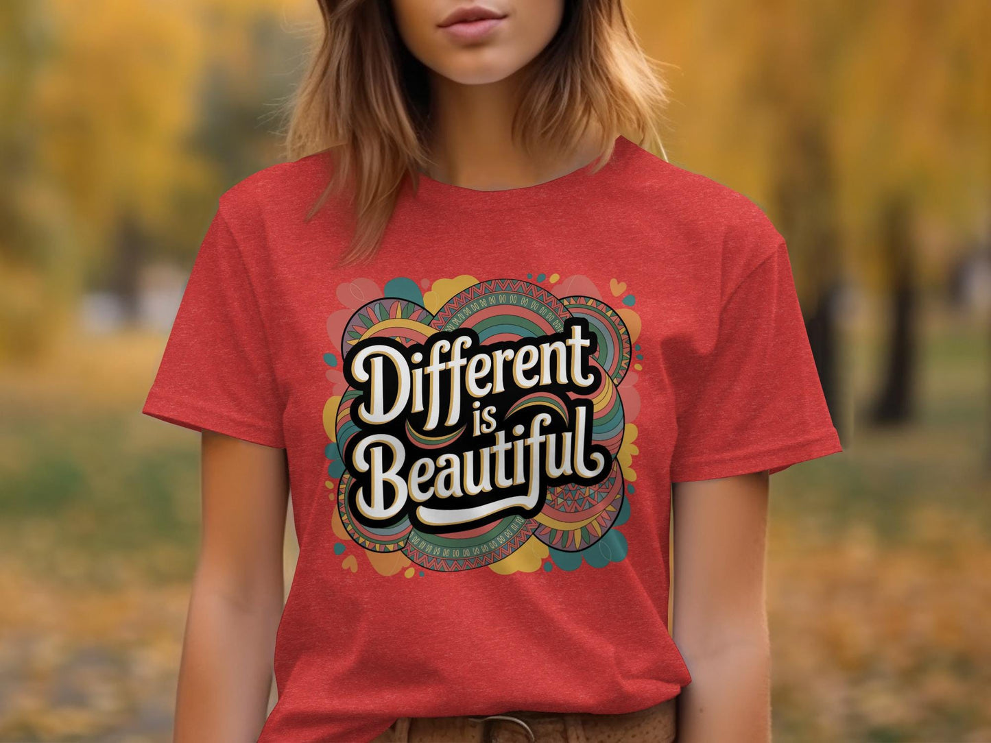 Woman wearing a red heather t-shirt with a bold retro-style graphic that says "Different is Beautiful" in large, white vintage lettering. The colorful background design features swirls, hearts, and abstract shapes in teal, mustard, orange, and pink. The shirt has a classic unisex fit with rolled sleeves, set in an outdoor autumn scene with yellow leaves in the background. A vibrant and inclusive fashion statement.