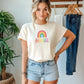 Stylish woman in a cream t-shirt with rainbow “BEAUTIFUL” text, wearing denim shorts and standing in a bright room with plants and hanging clothes.