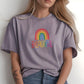 Woman in a soft lavender t-shirt with rainbow “BEAUTIFUL” print, sitting casually with visible floral tattoos and loose waves in hair.