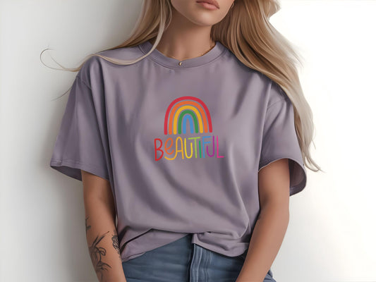 Woman in a soft lavender t-shirt with rainbow “BEAUTIFUL” print, sitting casually with visible floral tattoos and loose waves in hair.
