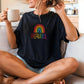 Woman relaxing on a couch wearing a black t-shirt with colorful “BEAUTIFUL” text and rainbow design, holding a mug and smiling.