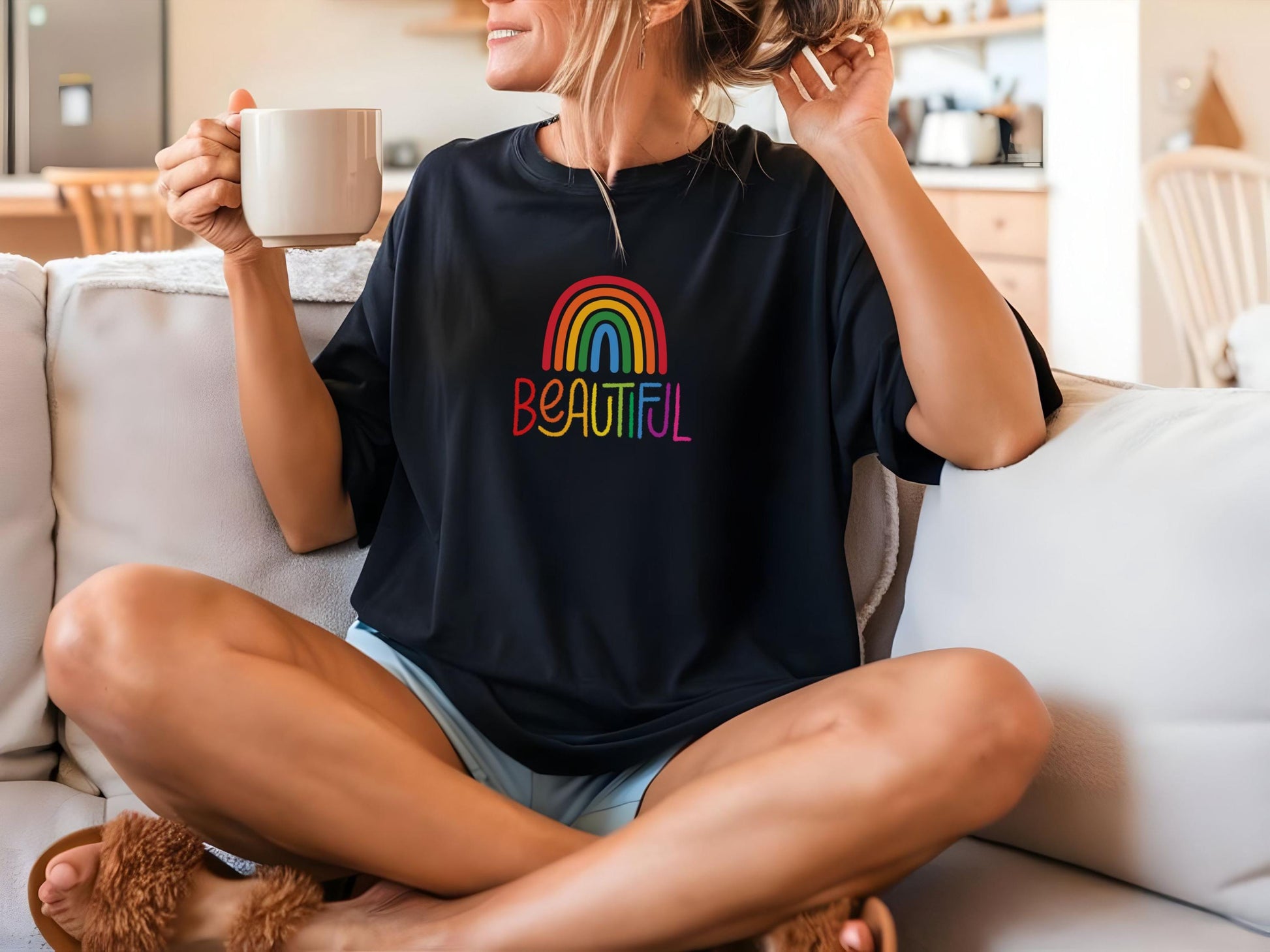 Woman relaxing on a couch wearing a black t-shirt with colorful “BEAUTIFUL” text and rainbow design, holding a mug and smiling.