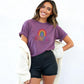 Smiling woman in a purple rolled-sleeve t-shirt featuring a rainbow and “BEAUTIFUL” design, paired with black shorts and a cozy cream cardigan.