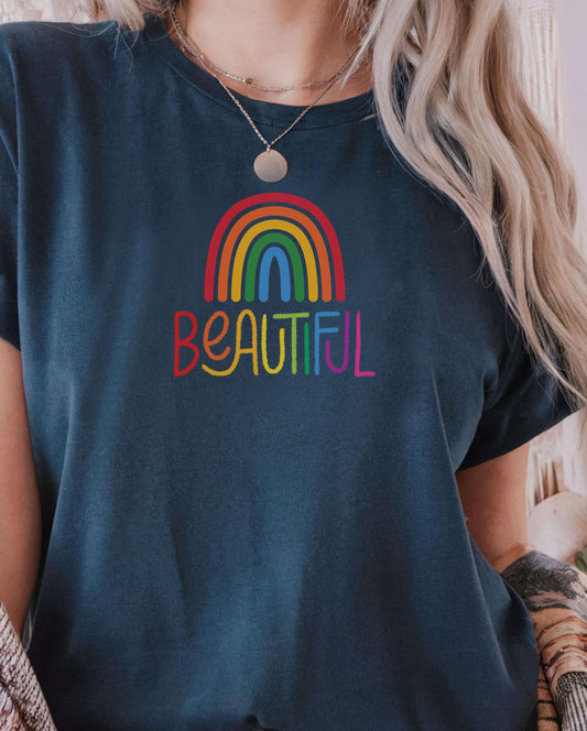 Woman wearing a dark blue t-shirt with a colorful rainbow design and the word “Beautiful” in rainbow lettering. LGBTQ+ pride shirt, positive message tee, inclusive fashion, casual uplifting apparel.