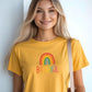 Blonde woman wearing a sunny yellow t-shirt with rainbow “BEAUTIFUL” graphic, smiling and holding a tote bag against a light wall.