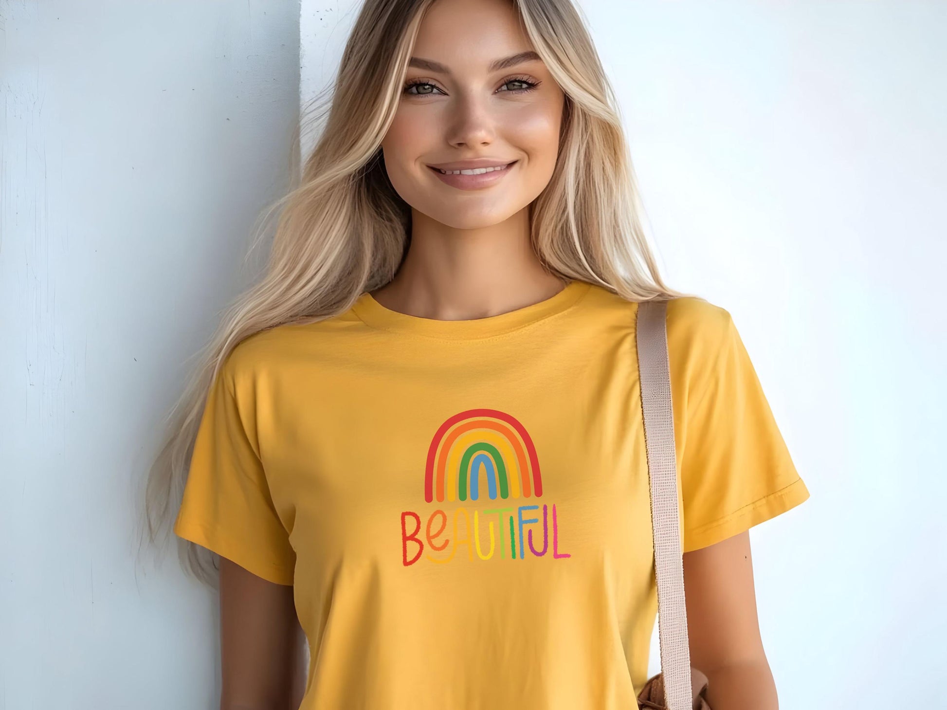 Blonde woman wearing a sunny yellow t-shirt with rainbow “BEAUTIFUL” graphic, smiling and holding a tote bag against a light wall.