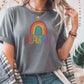 Woman in a soft gray t-shirt with rainbow “Beautiful” graphic, styled with layered necklaces and casual denim. Rainbow aesthetic shirt, modern inclusive tee, stylish mental health awareness shirt.