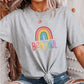 Light gray t-shirt with rainbow “Beautiful” text and rainbow graphic, styled with ripped jeans and a knotted front. Worn by a woman with wavy blonde hair and layered gold jewelry. LGBTQ+ pride shirt, positive message tee, rainbow graphic top, empowering casual outfit.