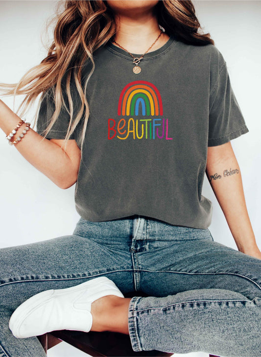 Woman sitting cross-legged on a stool wearing a faded charcoal t-shirt with a colorful rainbow and the word “BEAUTIFUL” printed in rainbow colors. She’s accessorized with a necklace, beaded bracelets, and casual jeans, creating a relaxed, boho-chic vibe.