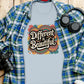 Flat lay of a light blue t-shirt featuring a bold retro graphic that says “Different is Beautiful” in large white lettering, surrounded by colorful swirls and hearts in teal, pink, orange, and yellow. Styled with a blue flannel shirt, headphones, and a camera. This colorful graphic tee is a positive quote tee, custom graphic print, and perfect as a stand out shirt, bold shirt, and self love t shirt. A unique statement tee and inspirational shirt for everyday wear.