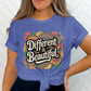 Woman wearing a tied royal blue t-shirt with a vibrant retro design that reads “Different is Beautiful” in large white lettering. The design includes colorful swirls, hearts, and patterns in pink, yellow, teal, and orange. This colorful graphic tee is a bold shirt, perfect as a positive quote tee, emotional shirt, and self love t shirt. A custom graphic print that works as a unique statement tee, colorful design tee, and inspirational shirt for those who love a stand out shirt.