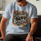 Man sitting indoors wearing a light blue t-shirt with a bold retro design that says “Different is Beautiful” in large white lettering. The colorful graphic includes hearts, swirls, and abstract patterns in teal, pink, mustard, and orange. This positive quote tee is a colorful graphic tee and custom graphic print, making it a bold shirt, emotional shirt, and unique statement tee. Perfect for anyone wanting a stand out shirt or self love t shirt with a positive message top.