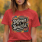 Woman wearing a red heather t-shirt with a bold retro-style graphic that says &quot;Different is Beautiful&quot; in large, white vintage lettering. The colorful background design features swirls, hearts, and abstract shapes in teal, mustard, orange, and pink. The shirt has a classic unisex fit with rolled sleeves, set in an outdoor autumn scene with yellow leaves in the background. A vibrant and inclusive fashion statement.