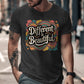 Man wearing a dark heather black t-shirt featuring a bold retro graphic that says &quot;Different is Beautiful&quot; in white vintage-style lettering. The colorful design includes swirling patterns and hearts in orange, teal, mustard, and pink. The shirt has a fitted cut with short sleeves, shown in an urban street setting. A stylish and empowering tee with inclusive, positive vibes.